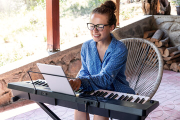 Young woman in glasses playing electric piano on terrace using digital tablet for online tutorial.