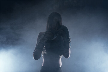 Dark contrast photo with smoke in the background of a focused strong woman with boxing gloves practicing punches