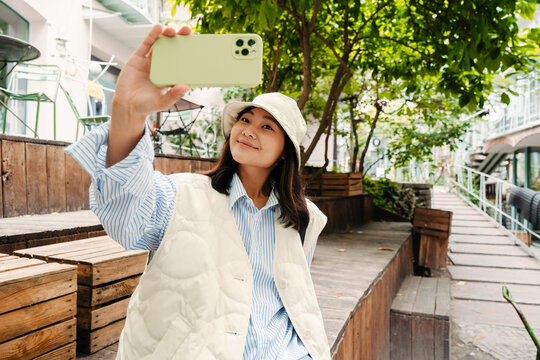 Smiling Asian Girl Taking Selfie With Mobile Phone While Sitting Outdoors