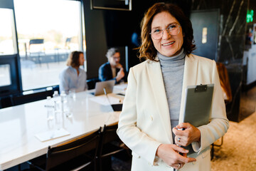 Mature woman in jacket smiling and holding clipboard during offline meeting
