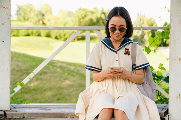Naklejka premium Young beautiful asian girl using mobile phone while sitting on bench in park