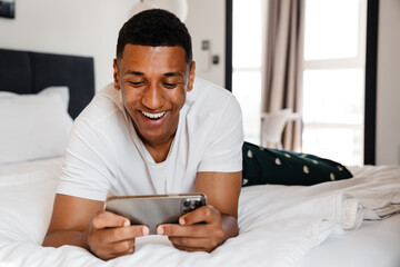 Excited african man playing game on smartphone while lying in bed