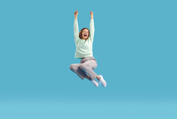 Joyful excited young woman laughing wildly, clenching her fists in joy of victory or success. Young woman screaming joyfully with closed eyes, raising hands and jumping high on light blue background.