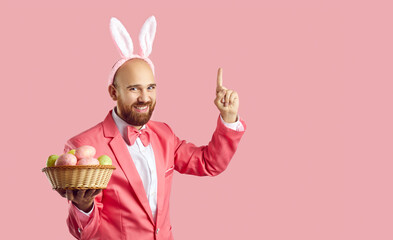Funny bald man with ginger beard, wearing suit, bow tie and cute bunny ears standing on pink background, holding bowl of Easter eggs, smiling, pointing first finger up, suggesting fun interesting idea