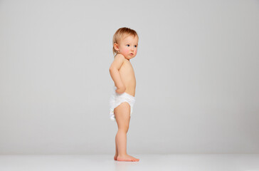 Little sad scared toddler girl, baby in diaper standing isolated on grey studio background. Concept...