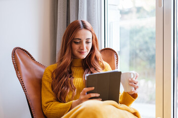 Young redhead woman reading at home with the tablet and having a coffee next to the window of her house