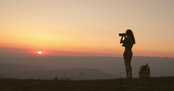 Dark silhouette of a slender young woman photographer at an orange summer sunset. Millennial creator woman takes beautiful pictures with a camera. Silhouette of a woman photographer at sunset. 