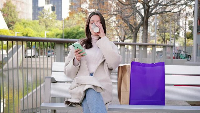 Happy young woman sitting on a bech using a smartphone to text messages after shopping on-line. Brunette female looking to the cellfone device sharing content on social media and drinking coffee