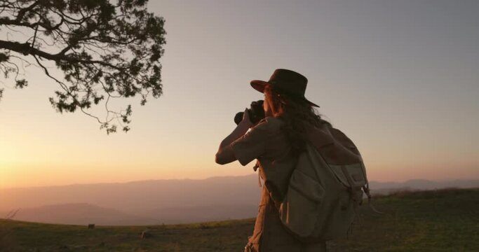 Female traveler photographer, illuminated by soft orange rays of the sun, enjoys a beautiful summer sunset in the mountains and takes epic pictures of setting sun. Silhouette of a woman photographer
