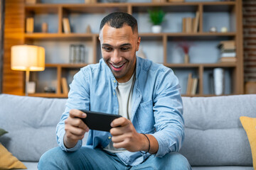 Joyful African American man is enjoying playing video game in smartphone having fun, rest sitting on sofa in apartment. Cool game with modern graphics and fun game play. People and gadgets concept.