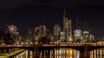 Fototapeta na wymiar New years eve with fireworks above the skyline of Frankfurt - Main at night at a cold day in winter with colorful reflections in the water.