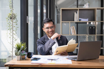 Fototapeta premium Young Asian businessman in suit and holding hot coffee cup and looking on laptop computer in office. Man in suit using laptop in well-lit workplace.