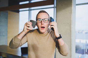 A cute young man in a brown sweater and glasses is talking emotionally on a mobile phone and drinking coffee while relaxing in the office. A young freelancer works remotely.