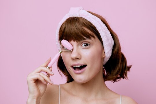 A Beauty Photo Of A Funny, Shocked Woman With Short Hair Tucked Behind A Soft Headband And Holding A Pink Facial Massager In Her Hand Covers Her Face With It From The Camera