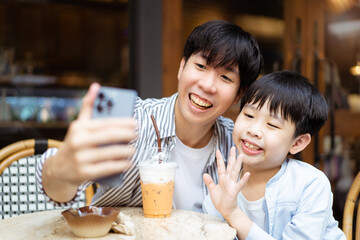 Happy Asian father and son enjoy a selfie photography or video call with their family and friends together while traveling. Cheerful young boy taking and taking a photo on smartphone with his dad.
