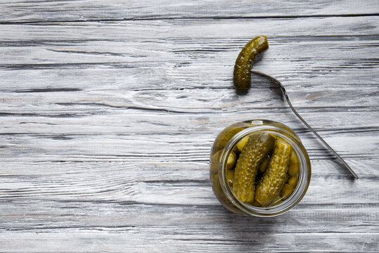 Open jar with pickled cucumbers and cucumber on a fork on a light wooden background top view.