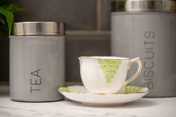 selected focus on bone china cup and saucer with tea and biscuit cannisters in the background on a marble work top