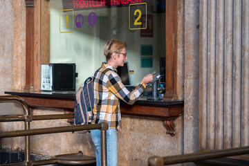 Adult 35s year old lesbian woman in plain shirt and jeans with backpack and sunglasses traveling by train in Europe. Pay with bank card for the ticket on train station in Barcelona, Spain.