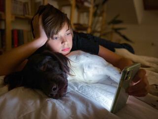 Boy with looking at smart phone lying of the bed in the room with his dog. Copy space, miejsce na...
