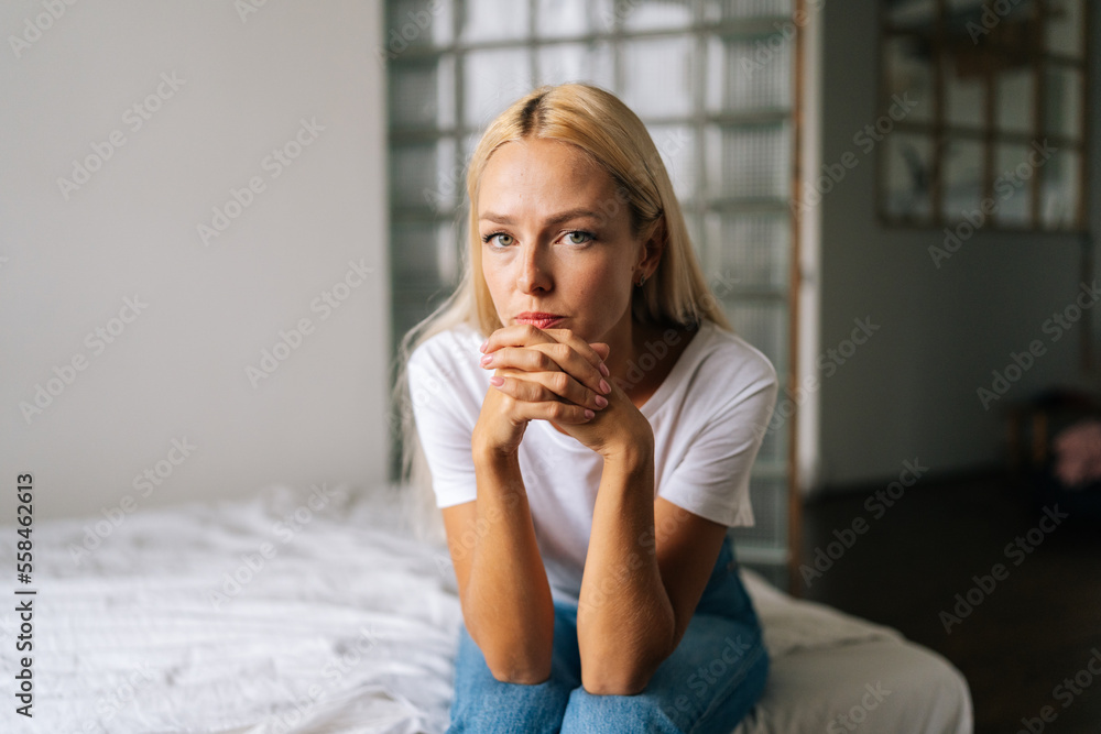 Wall mural close-up portrait of pensive young blonde woman sitting alone at in living room and sad looking came