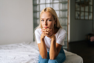 Close-up portrait of pensive young blonde woman sitting alone at in living room and sad looking camera holding hands on chin, thinking over health problems, feeling sadness, boredom, apathy.