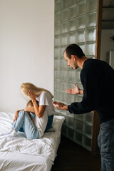 Vertical shot of scared mother and daughter hugging each other sitting on sofa while aggressive father angrily yelling at them at home. Drunken husband scolding, raising voice, scream at family