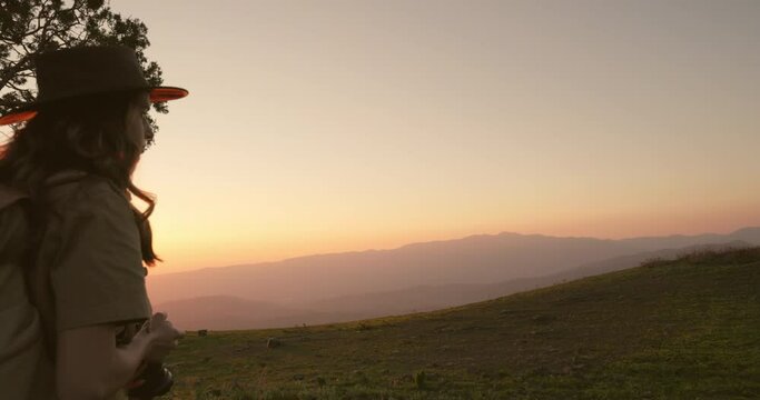 Shot from behind woman traveler with hiking backpack, in safari outfit on top of hill looking for best place to take beautiful sunset shots in mountains. Red-orange sunset in hilly, mountainous area.