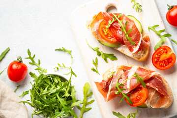 Open sandwiches with cream cheese, prosciutto, cucumber and arugula at white table.