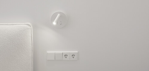 Bedside white LED wall lamp illuminating. Creative modern wall light source in bedroom. White European electrical socket outlet and switch. Electricity and illumination concept.