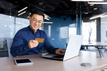 Happy and successful businessman in office making online payments inside building, asian man using bank credit card and laptop for online shopping, man working sitting at desk.