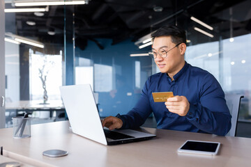 Happy and successful businessman in office making online payments inside building, asian man using bank credit card and laptop for online shopping, man working sitting at desk.