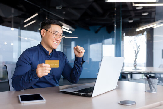 Happy And Successful Businessman In Office Making Online Payments Inside Building, Asian Man Using Bank Credit Card And Laptop For Online Shopping, Man Working Sitting At Desk.