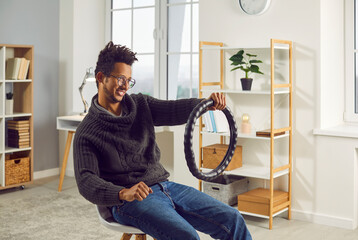 Funny, happy young African American man sitting on a chair in the living room at home, holding a...