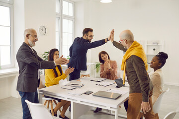 Two happy and satisfied male colleagues give each other high five during business meeting. Business people sitting at table applaud congratulating their colleagues on job well done and successful deal - obrazy, fototapety, plakaty