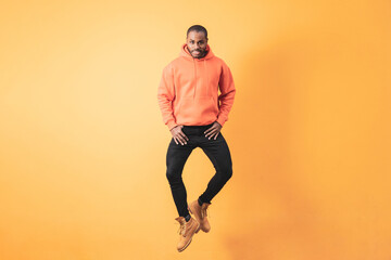 Fototapeta na wymiar A dark-skinned man in an orange hoodie looks at the camera as he jumps in front of an orange wall.