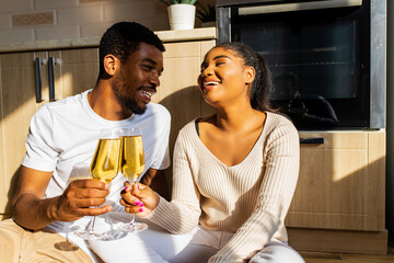 Laughing young afro ethnic couple dating with champagne at home while sitting at the warm floor