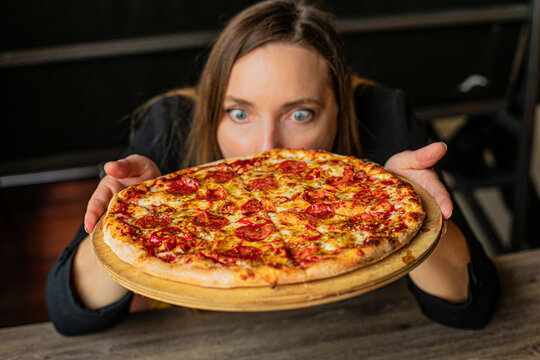 Hungry woman hold in hand wooden tray with fresh appetizing pizza and gawp. Serving of hot flavored pepperoni with crisp crust to guests. Italian kitchen, pizzeria restaurant, dinner, flour food