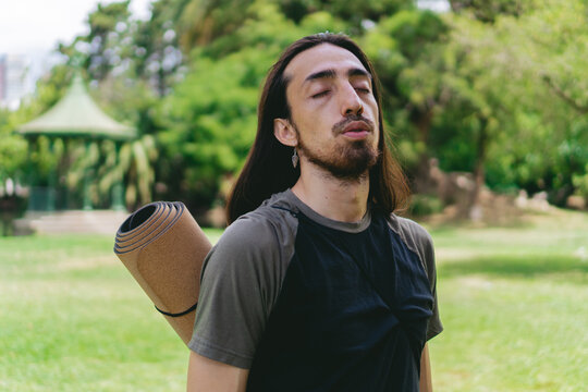 Young, Latino, Hispanic Hippie Male With Long Hair Breathing Deeply In A Natural Environment With A Gazebo Behind Him.