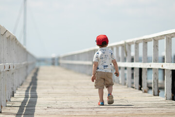 little boy walking alone on a pier wearing baseball hat on summer day