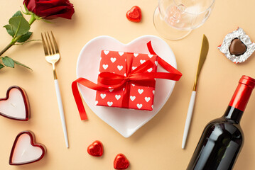 Valentine's Day concept. Top view photo of heart shaped plate with giftbox knife fork wine bottle candles glass chocolate candies and rose on isolated pastel beige background