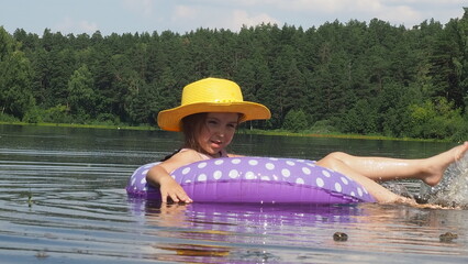 Girl in a hat swims on the river bank or lake with a inflatable circle. Splashes water with feet. Hot summer. Beach holidays, swimming, tanning, sunscreens.