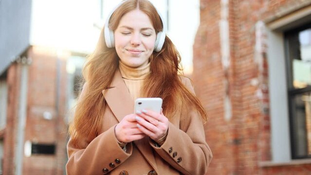 Portrait of smiling red haired ginger young woman with headphones listening to music while hold smartphone scrolling social media texting browsing online checking email on the brick street alone