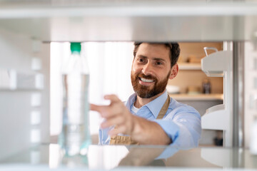 Smiling millennial caucasian bearded guy in apron opens door of empty refrigerator and takes bottle of water