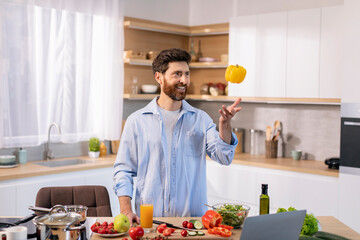 Cheerful millennial caucasian guy cooks eat, catches pepper, has fun at table with vegetables and...