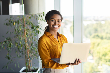Portrait of happy businesswoman using laptop in office interior, standing near window and smiling...