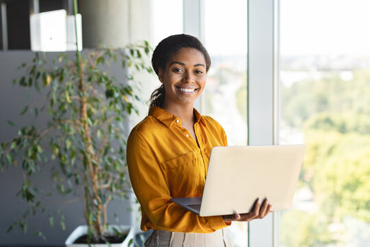 Portrait of happy businesswoman using laptop in office interior, standing near window and smiling at camera