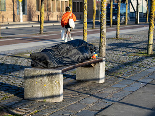 Homeless person sleeps on a bench in a park in dark and dirty sleeping bag woman out of focus...
