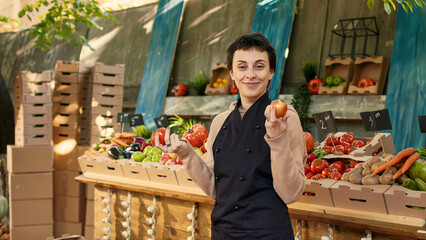 Farmers market vendor showing organic produce to clients, presenting colorful fruits and vegetables on food marketplace stand. Young adult having farming counter to sell products.