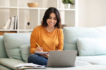 Smiling Young Arab Female Student Using Laptop At Home And Taking Notes