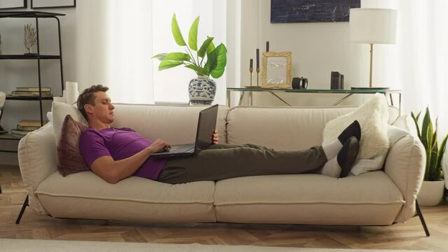 A man works with a computer at home while lying on the sofa. Comfortable remote work online. A freelancer performs tasks. Handsome Caucasian young man working on laptop. High quality 4k footage